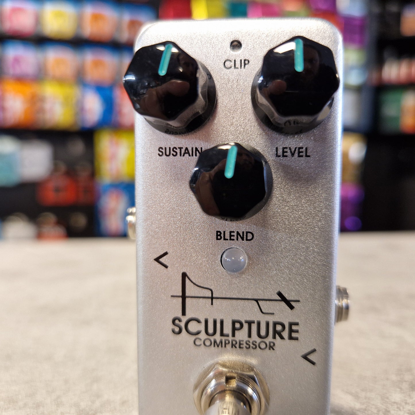 Guitar effects pedal with 'Sculpture Compressor' branding on a blurred store background.
