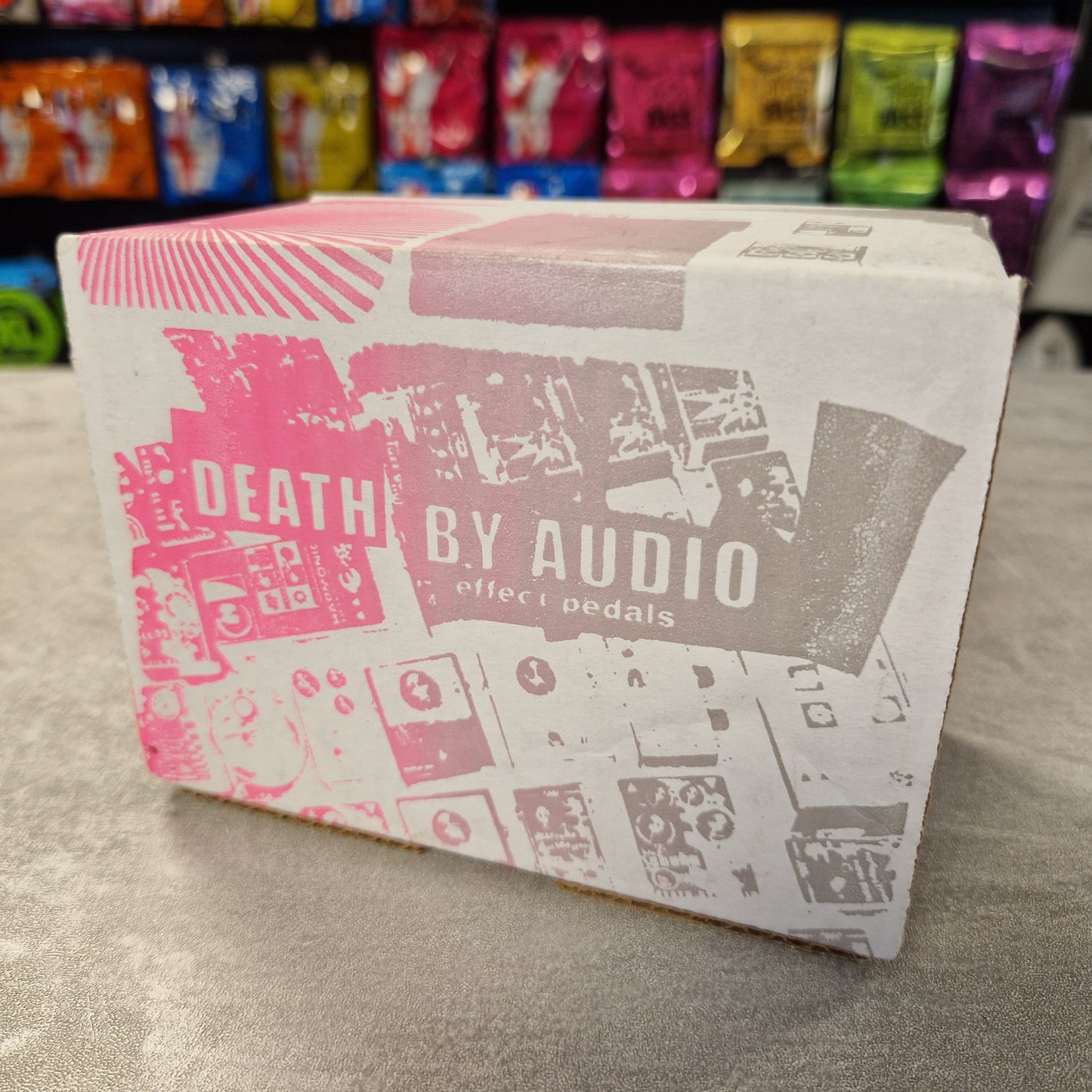 Pre-Owned Death by Audio Box with 'Death by Audio' branding on a gray surface with colorful packaging in the background.