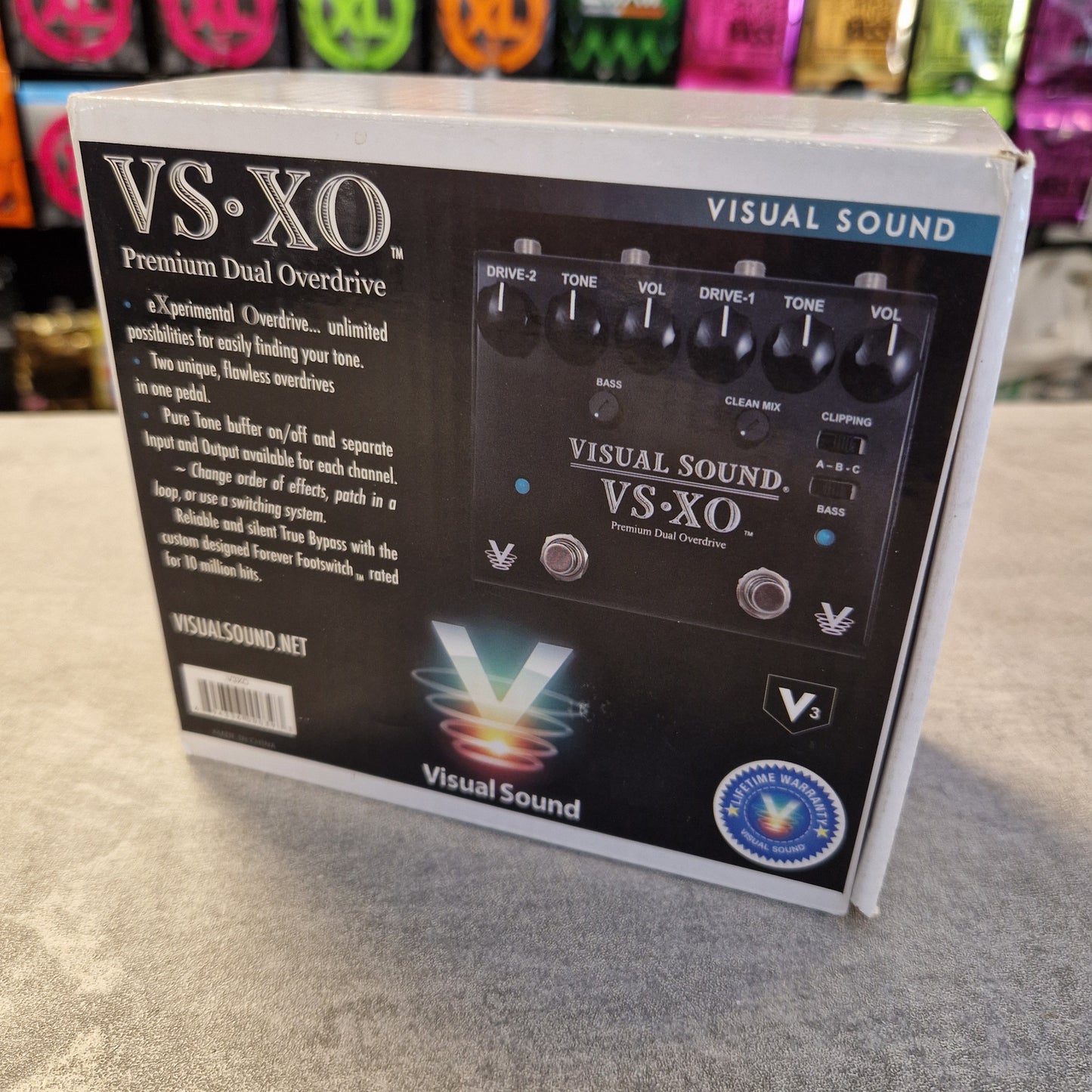 Pre-Owned Truetone VSXO Visual Sound VS:XO guitar effects pedal packaging on a gray surface with colorful pedals in the background.