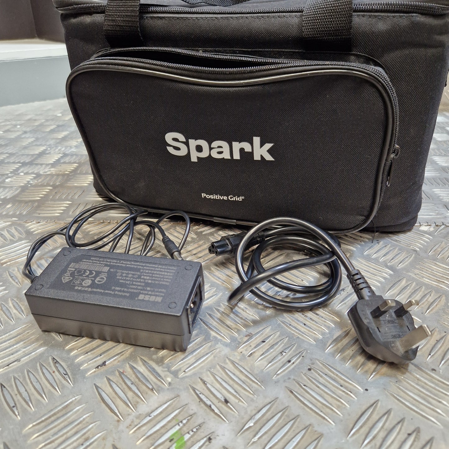 Pre-Owned Positive Grid Spark 40-Watt Smart Guitar Combo & Bluetooth Speaker A black Spark Positive Color portable light bag shown with its power adapter and UK plug on a diamond plate metal surface.