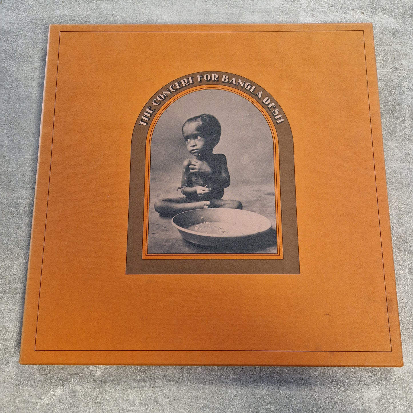 Various - The Concert For Bangla Desh Vintage Vinyl Boxset A square orange album cover or book-style package photographed on a gray textured surface. The center features an arched inset with a black-and-white photo of a child and the text 'HE'S GOT TO BE A MAD-ASS' around the image.
