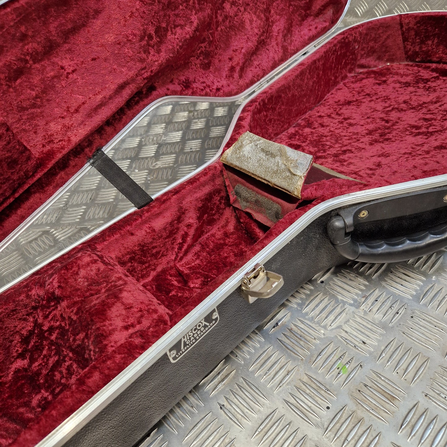 Pre-Owned Hiscox LiteFlite Acoustic Hard Case A close-up of an open hard instrument case with deep red velvet lining and a black exterior shell, photographed on a metal diamond-plate surface.