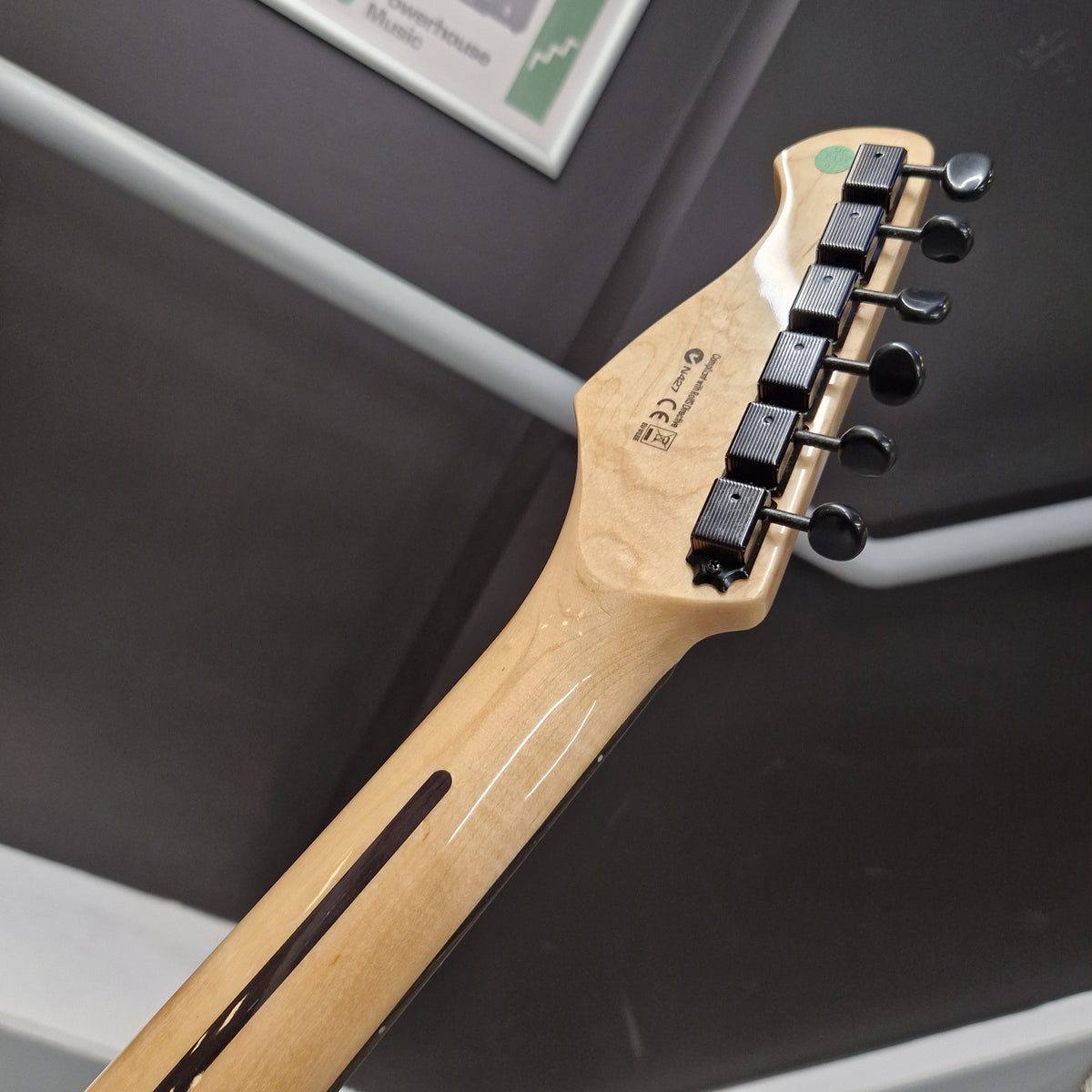 Pre-Owned Harley Benton JA-Baritone HH Electric - Black Close-up of the headstock of a guitar with a natural light wood finish and black tuning pegs, photographed indoors against a dark gray background.