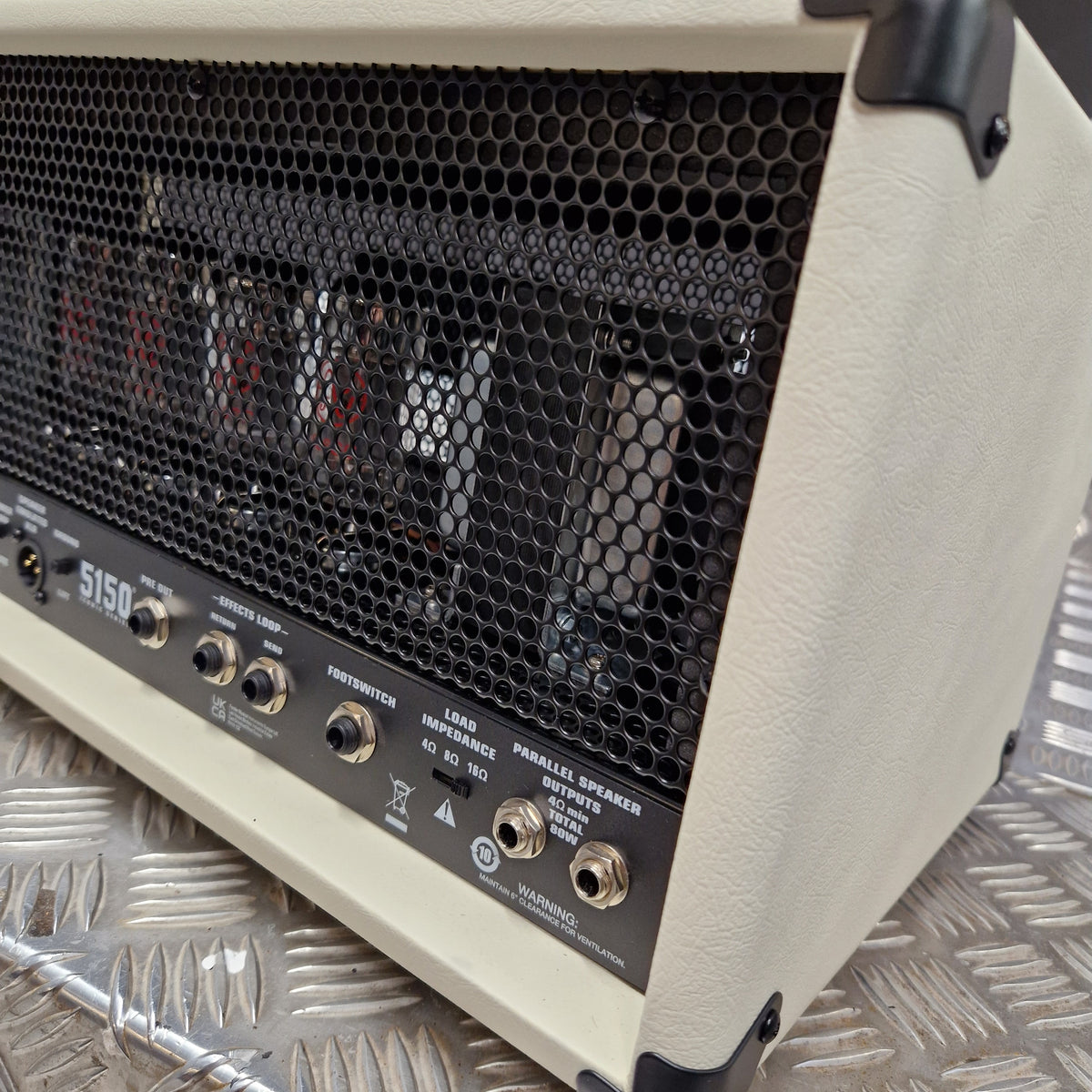 Pre-Owned EVH 5150 Iconic 80W Electric Amplifier Head Close-up of the rear corner of a cream-colored Marshall 5150 guitar amplifier head, showing the black honeycomb grille, control knobs, and internal components behind the grille, resting on a diamond plate metal surface.