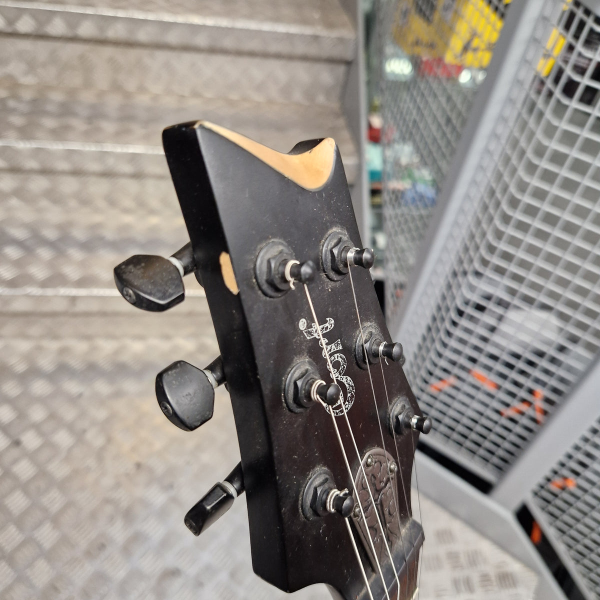 Pre-Owned Cort EVL-Z4 Electric - Satin Black Close-up of a black guitar headstock with four tuning pegs and four strings, photographed in a real industrial setting with metal steps and a mesh gate visible in the background.