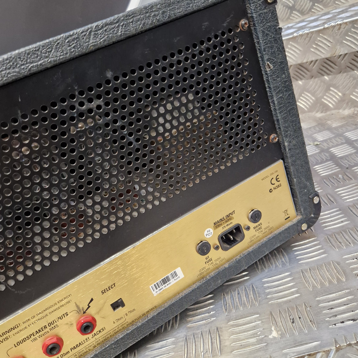 Pre-Owned Marshall JCM2000 DSL100 Valve Electric Amplifier Head Close-up of the rear panel of a guitar amplifier, showing the gold-colored backplate with input jacks, power switch, and speaker grille on a diamond plate background.