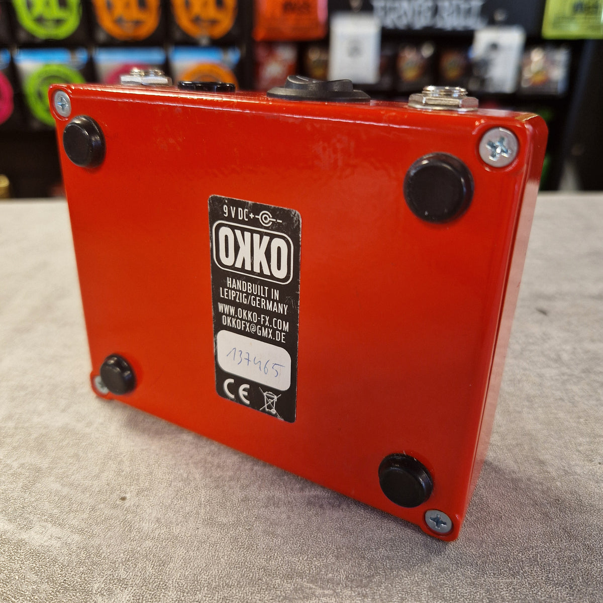 Pre-Owned Okko Dominator High Gain Distortion Pedal - An orange-red OXKO guitar effects pedal shown from an angled top-down view, resting on a textured gray surface with a blurred store background.