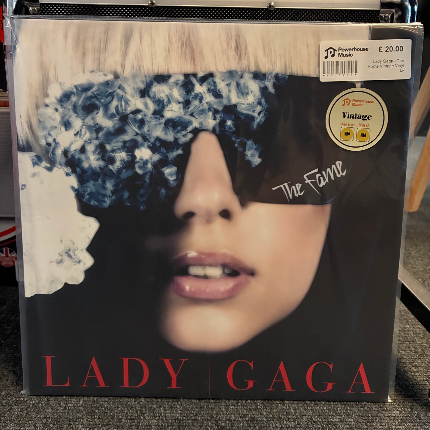 Lady Gaga - The Fame Vintage Vinyl LP A vinyl record sleeve of Lady Gaga's album 'The Fame' in its clear plastic shrink-wrap, resting on a gray carpeted shelf. The cover art features a close-up of a woman's face with a blue-and-white patterned blindfold and the title 'LADY GAGA' printed in red at the bottom.