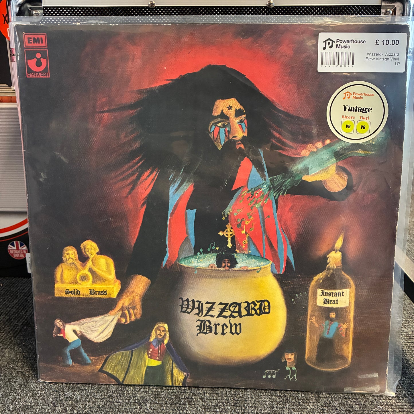 Wizzard - Wizzard Brew Vintage Vinyl LP Jazz Brew vinyl record album cover in a clear plastic protective sleeve, photographed standing on a gray carpet. The cover art shows a dark-haired figure in front of a cauldron with the title 'Jazz Brew' and smaller illustrated scenes around it.