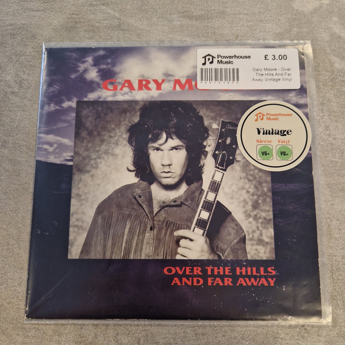 Gary Moore - Over The Hills And Far Away Vintage Vinyl 7" Single A vinyl record sleeve for Gary Moore's 'Over the Hills, and Far Away' on Powerhouse Music, sealed in a plastic bag with a price sticker and a vintage rating sticker on a gray textured surface.