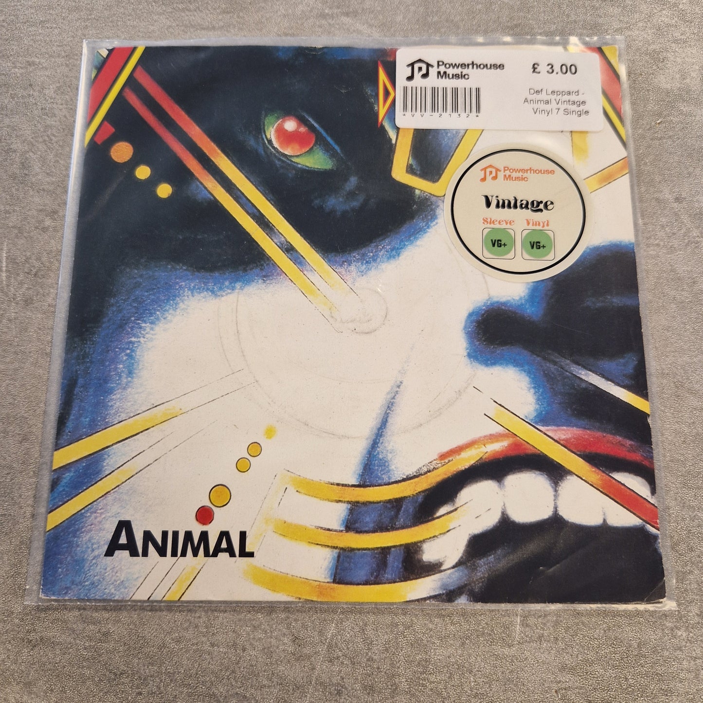 Def Leppard - Animal Vintage Vinyl 7" Single A square printed item, likely a sticker, card, or small art print, wrapped in clear plastic on a gray fabric surface. The artwork shows a stylized close-up face with blue, white, black, yellow, red, and orange geometric lines.