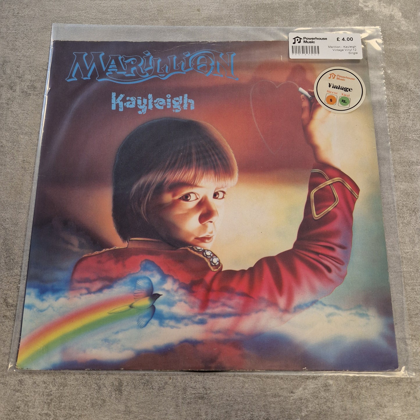 Marillion - Kayleigh Vintage Vinyl 12" Single A vinyl record sleeve for Marillion's 'Kayjish' is sealed in a clear plastic sleeve and photographed on a gray textured surface. The cover art features a surreal portrait of a child in a red jacket against a sky-like background with a rainbow.