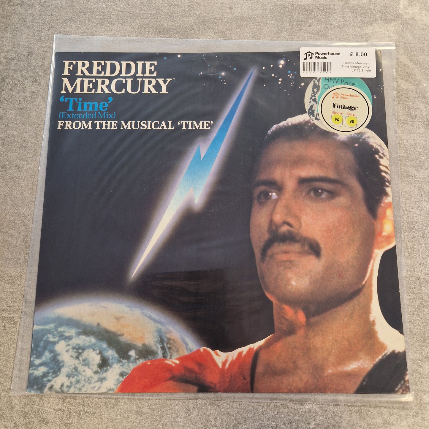 Freddie Mercury - Time Vintage Vinyl LP 12" Single Freddie Mercury's 'Time' vinyl record album cover, sealed in a clear plastic sleeve, displayed on a gray textured surface. The cover art features a close-up portrait of Freddie Mercury, a blue lightning bolt, and a partial view of Earth.