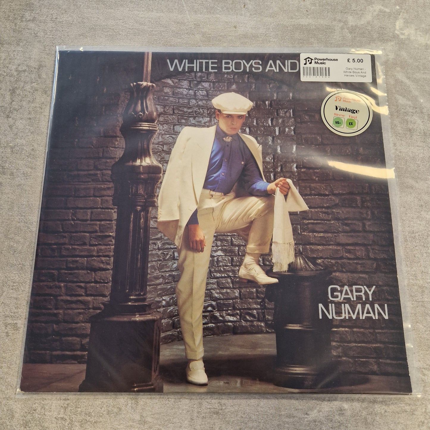Gary Numan - White Boys And Heroes Vintage Vinyl 12" Single A vinyl record sleeve for Gary Numan's 'White Boys and Black Girls' is sealed in plastic and photographed on a gray textured surface. The cover art features a man in a white blazer and beige trousers posing against a dark brick wall.