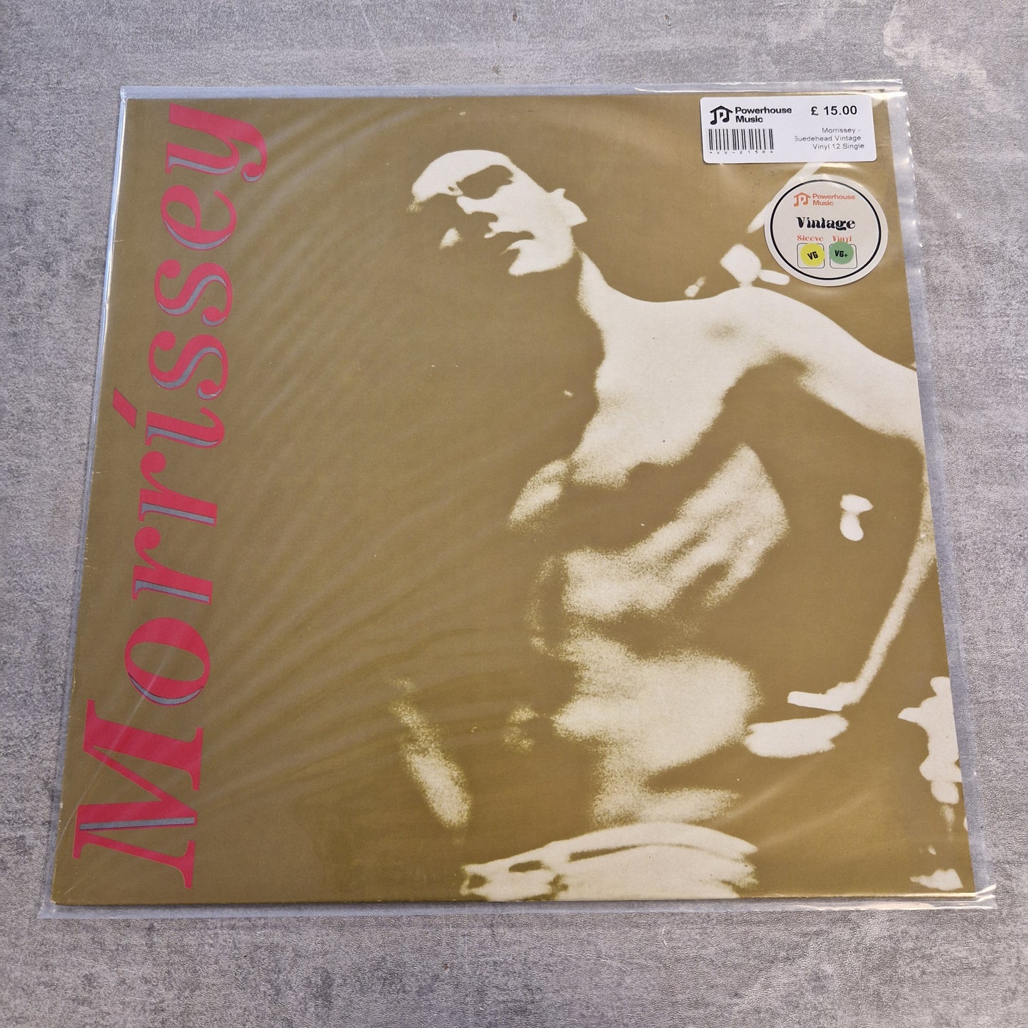 Morrissey - Suedehead Vintage Vinyl 12" Single A Morrissey vinyl record sleeve inside a clear protective plastic sleeve, placed on a gray textured surface. The cover features a sepia-toned abstract portrait of a shirtless figure with the name 'Morrissey' printed vertically in pink on the left side.