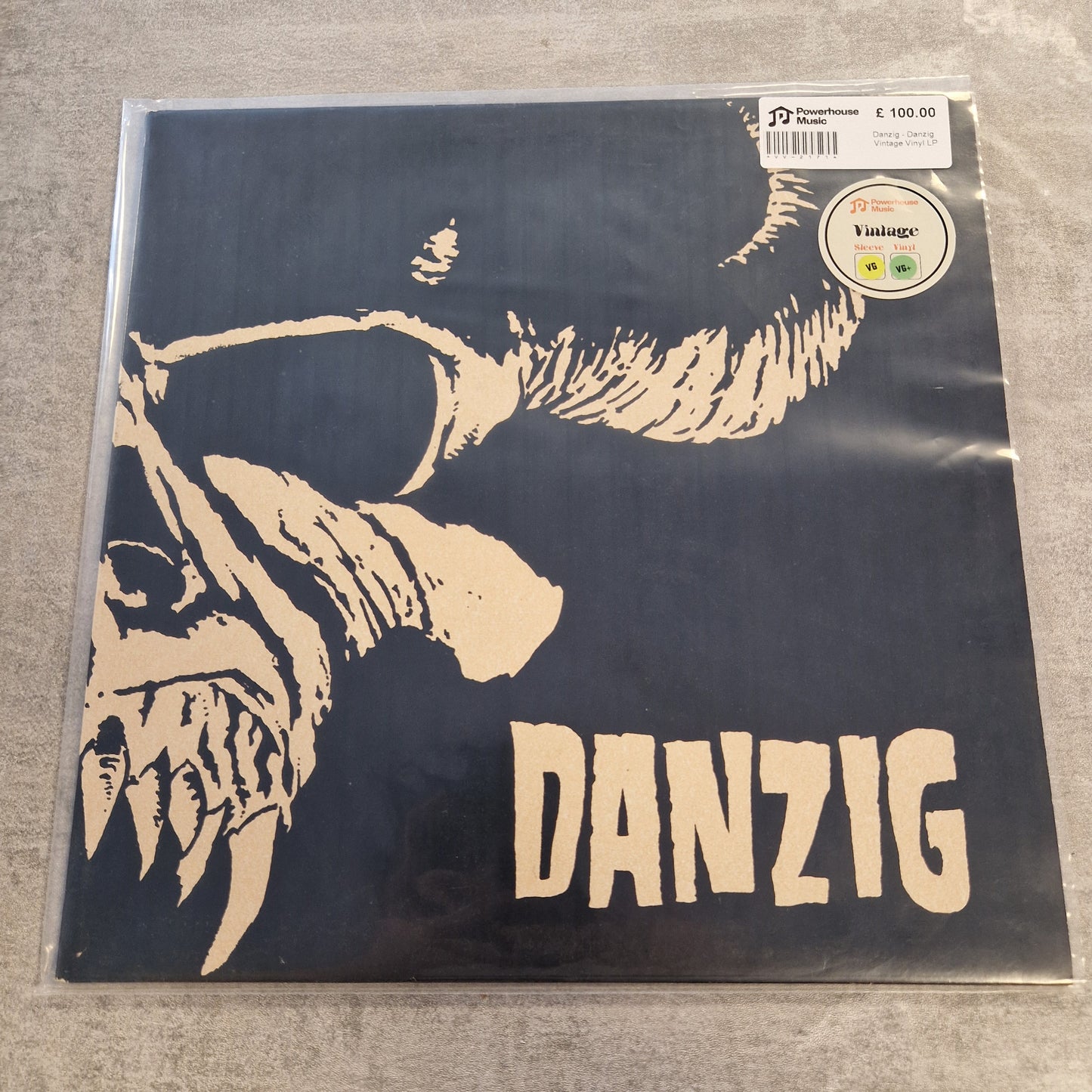 Danzig - Danzig Vintage Vinyl LP A vinyl record sleeve for the band Danzig, featuring a bold cream-colored graphic of a horned figure on a dark navy background. The record is sealed in a protective plastic sleeve, with a barcode sticker and a vintage label visible in the top right corner, placed on a gray textured surface.