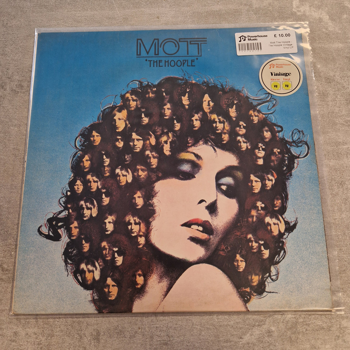 Mott The Hoople - The Hoople Vintage Vinyl LP Mott The Hoople 'The Hoople' vinyl record album cover in its original shrink wrap, displayed on a gray textured surface. The cover art features a striking portrait of a woman with voluminous dark curly hair composed of repeated faces, set against a bright blue background.