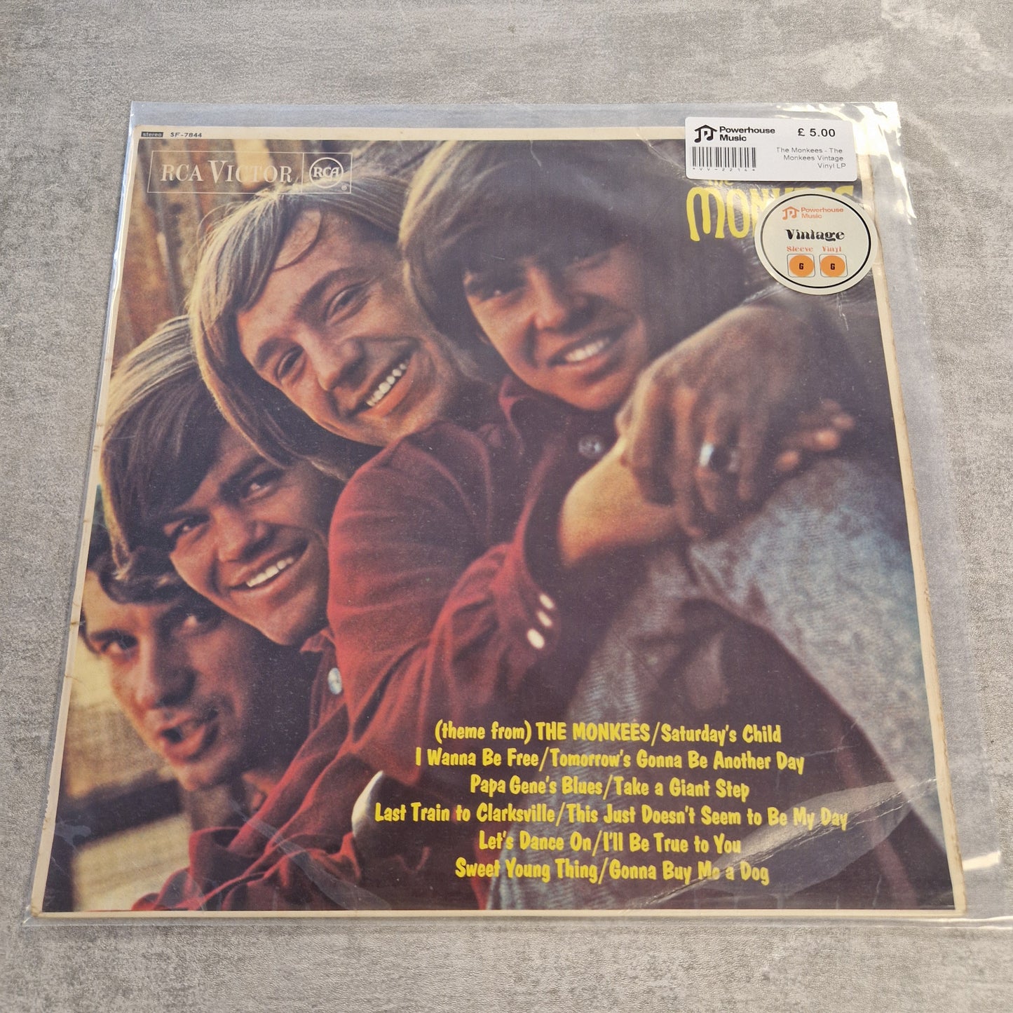 The Monkees - The Monkees Vintage Vinyl LP A vinyl record sleeve for RCA Victor's 'Names from The Monkees / Saturday's Child' album, sealed in plastic packaging and photographed on a gray textured surface. The cover features the four Monkees smiling closely together, with tracklist text in yellow at the bottom right.