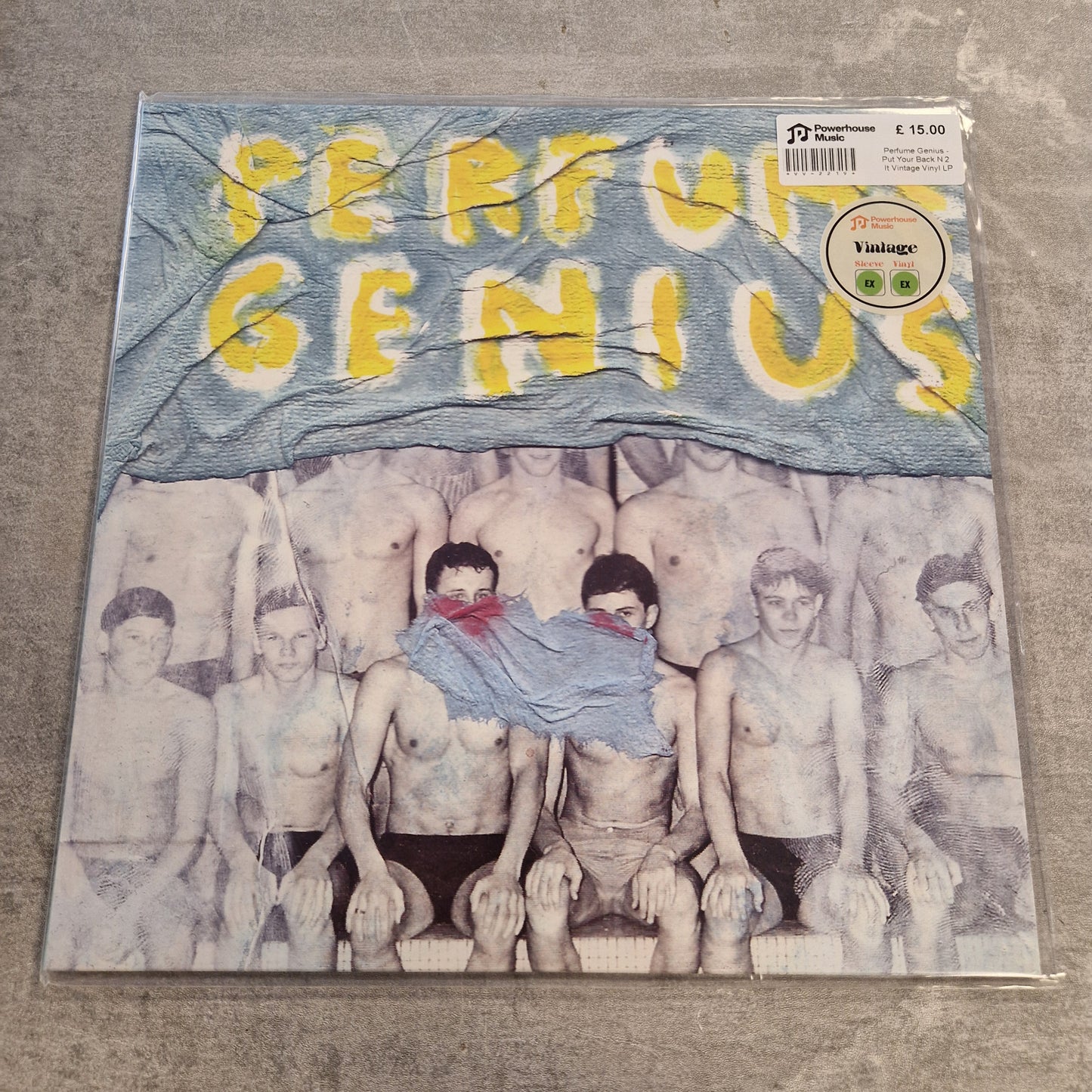 Perfume Genius - Put Your Back N 2 It Vintage Vinyl LP A vinyl record sleeve for the band 'Perfume Genius' lies on a gray concrete floor. The cover art features a blue flag with the band's name in yellow letters above a black-and-white photo of shirtless men.