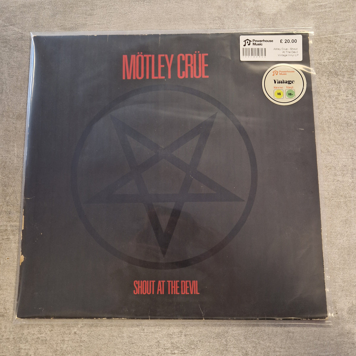 Motley Crue - Shout At The Devil Vintage Vinyl LP Mötley Crüe 'Shout at the Devil' vinyl record cover in its clear plastic shrink wrap, displayed on a gray textured surface. The cover is black with a large gray pentagram design and red text.