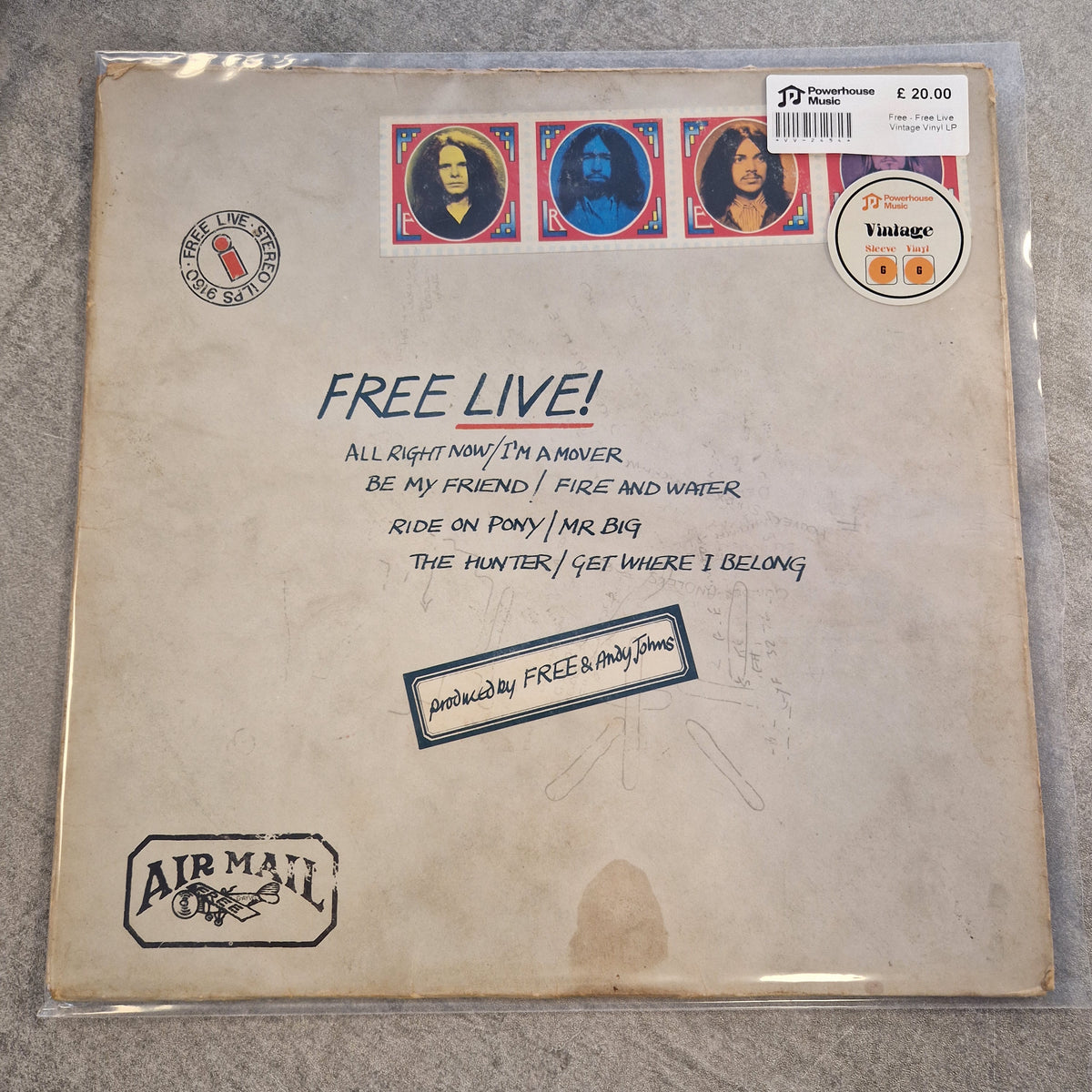 Free - Free Live Vintage Vinyl LP A vinyl record sleeve for Airmani's 'Free Live!' album, sealed in a clear plastic protective case and photographed on a gray textured surface. The sleeve features a vintage, off-white background with printed black text, postage stamp graphics on the left, and the Airmani logo in the bottom right corner.