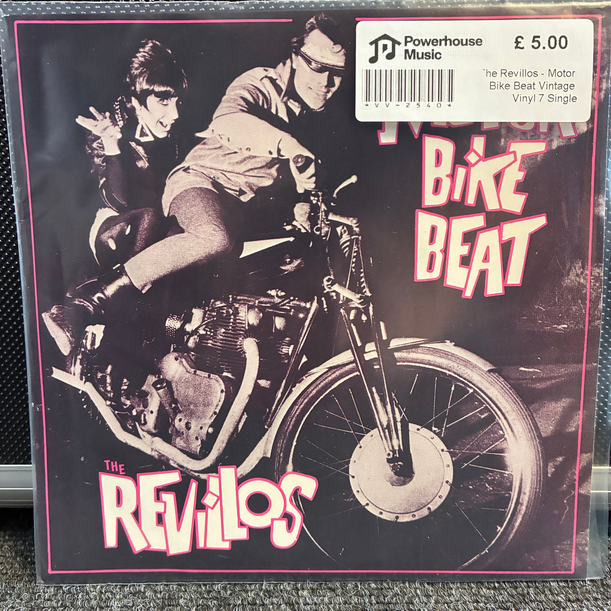 The Revillos - Motor Bike Beat Vintage Vinyl 7" Single Vinyl record single cover for 'Bike Beat' by The Revillos, featuring a black-and-white photo of two people riding a motorcycle with bold pink-outlined white text on a dark background. A Powerhouse Music price sticker is visible in the top right corner.