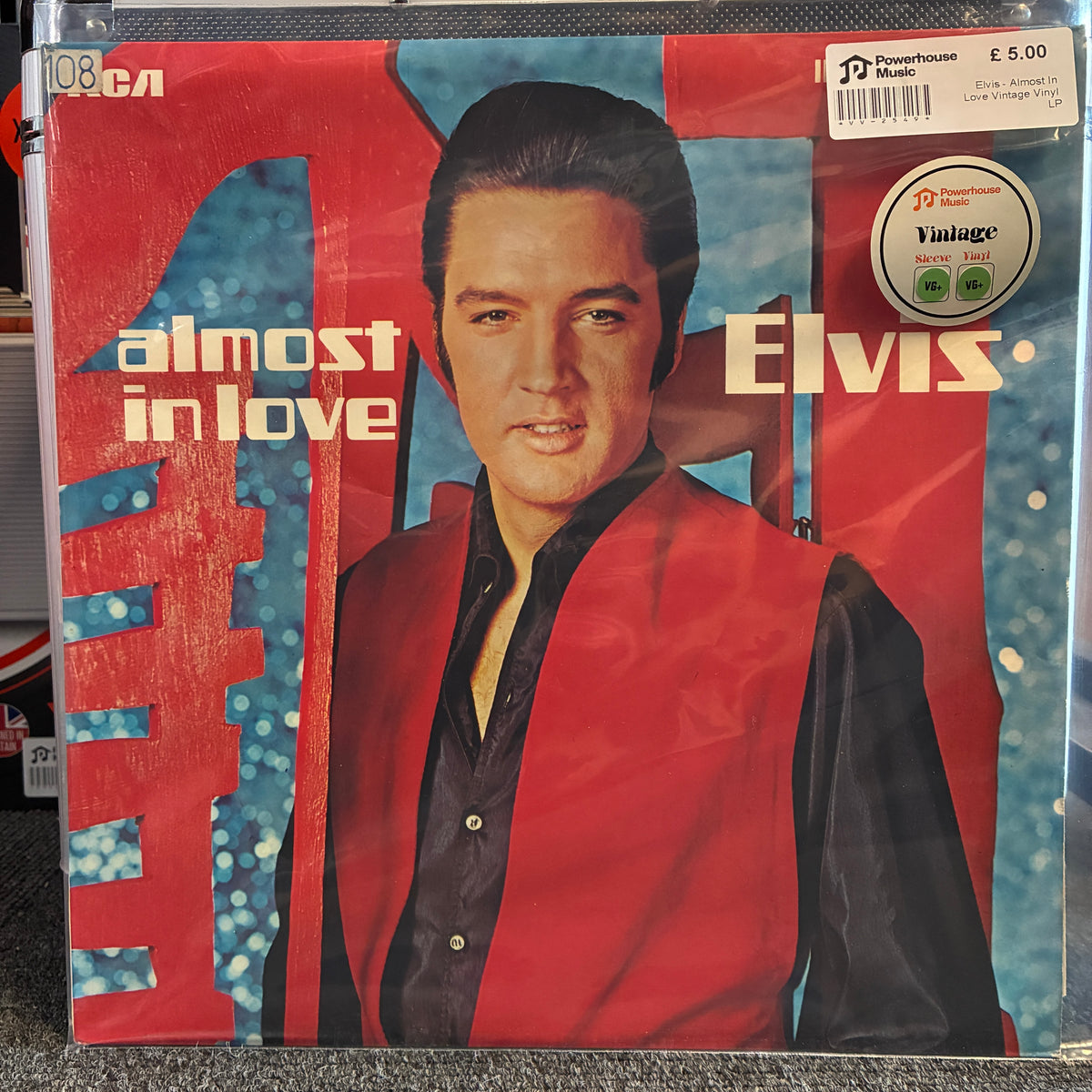 Elvis - Almost In Love Vintage Vinyl LP Elvis 'Almost in Love' vinyl record album cover, showing Elvis in a red vest over a black shirt against a blue bokeh background, with a visible Discogs sticker and a price tag on the cover.