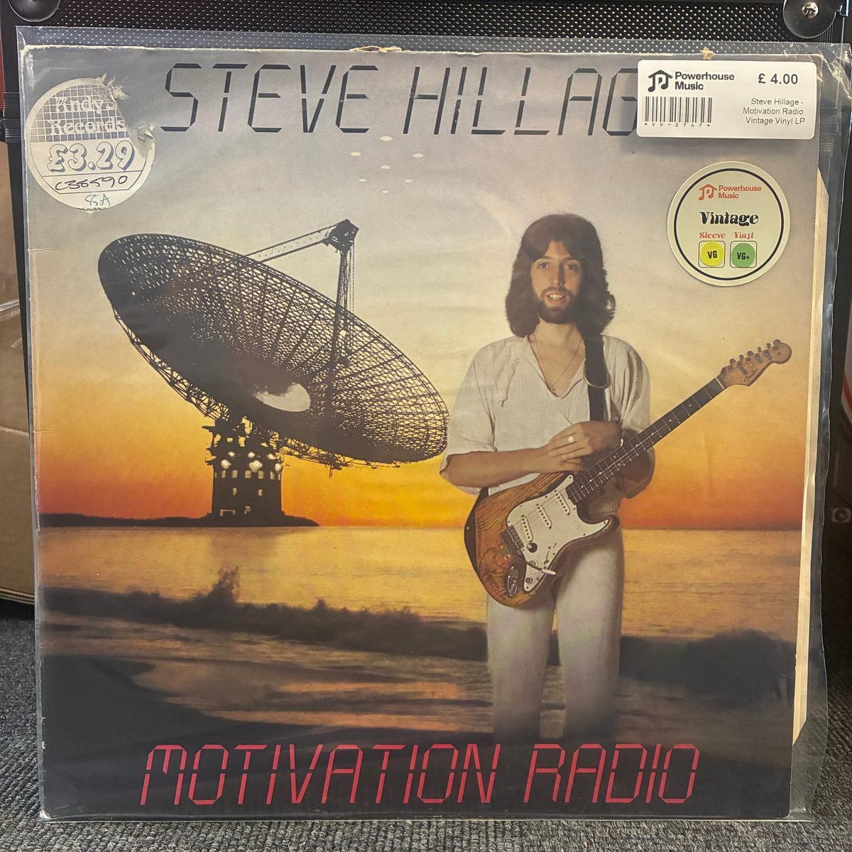 Steve Hillage - Motivation Radio Vintage Vinyl LP Steve Hillage 'Motivation Radio' vinyl record sleeve, sealed in a clear plastic protective case. The cover art features a sunset landscape with a large radio dish and a portrait of Steve Hillage holding an electric guitar.