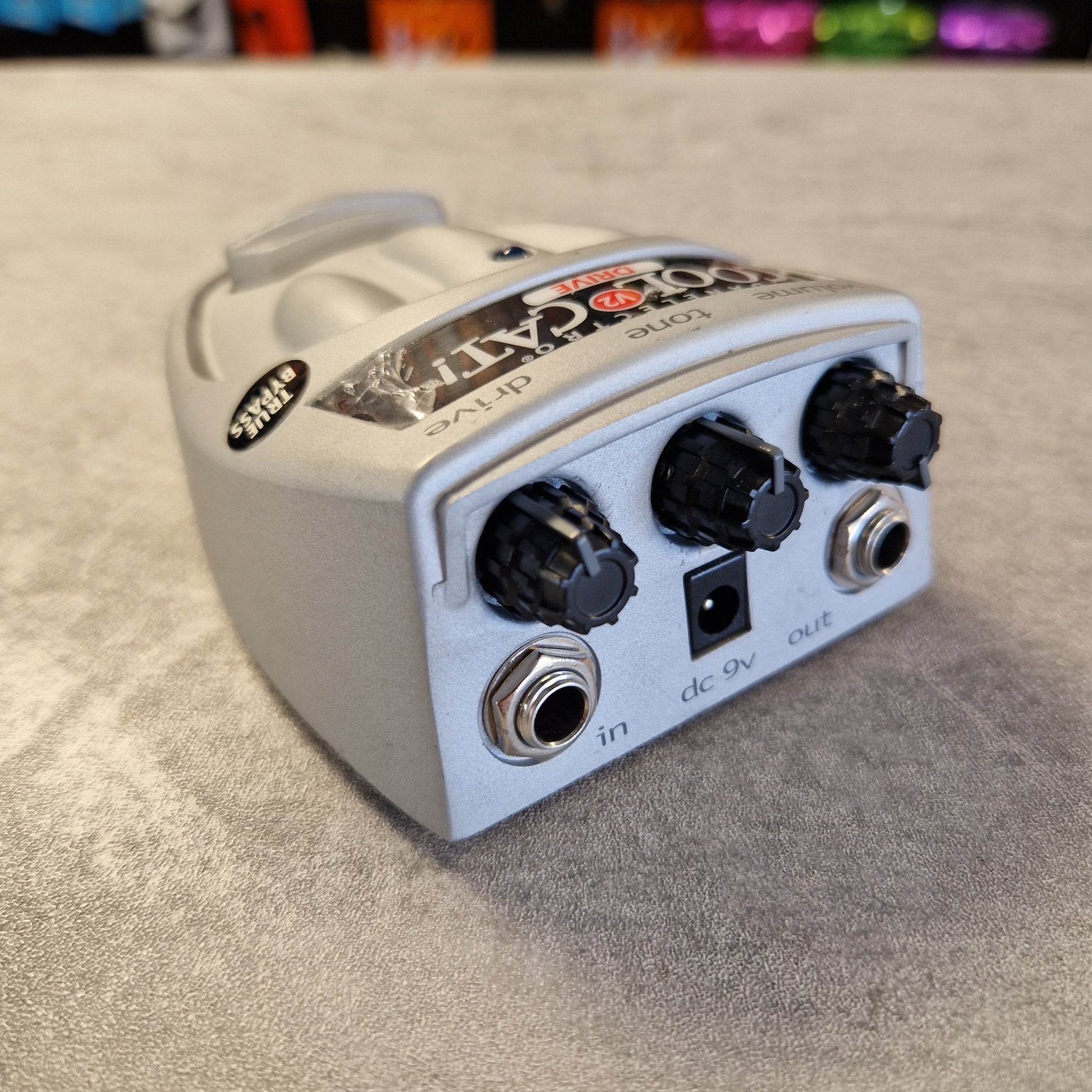 Danelectro Cool Cat V2 Drive Guitar effects pedal on a textured surface with colorful equipment partially visible.