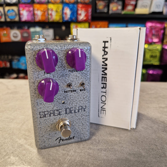 HammerTone Space Delay guitar effects pedal on a gray surface with colorful packaging in the background.