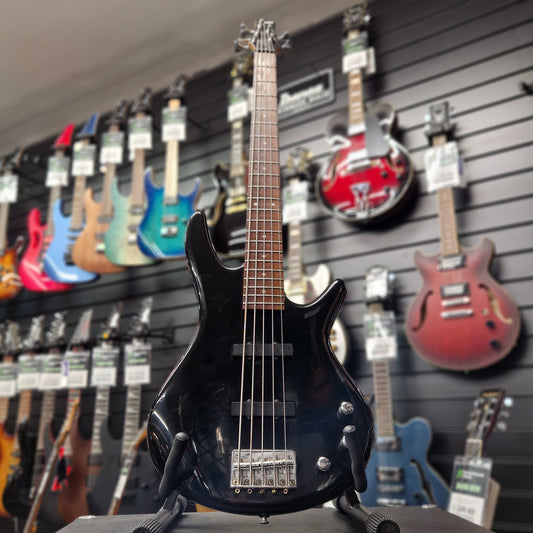 Pre-Owned Ibanez GSR205 Black electric guitar held in front of a wall of colourful guitars