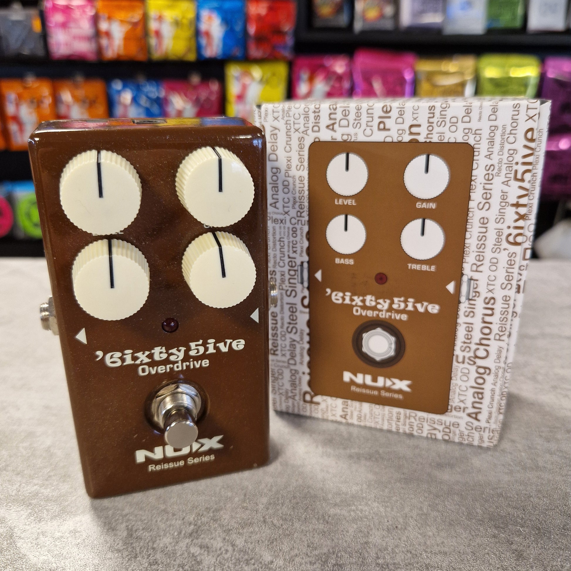Pre-Owned NUX guitar effect pedals on a grey surface with colourful boxes in the background.