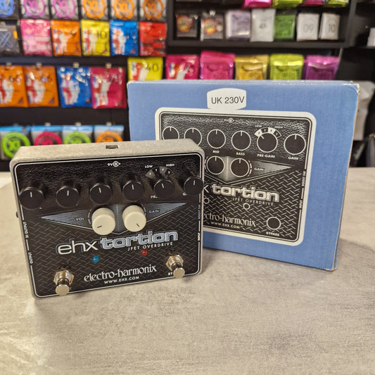 Pre-Owned Electro-Harmonix Two electronic effect pedals on a grey surface with colourful packaging in the background.