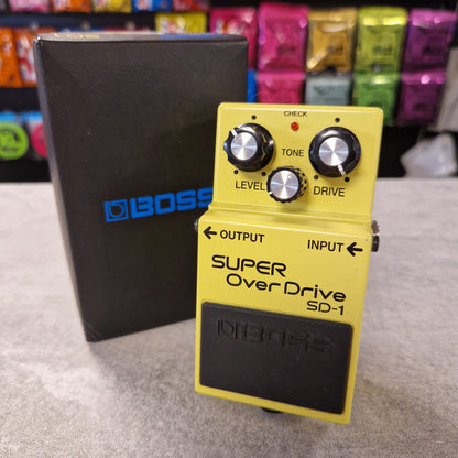 Pre-Owned Boss Yellow Boss Super Overdrive SD-1 pedal on a grey surface with colourful boxes in the background.
