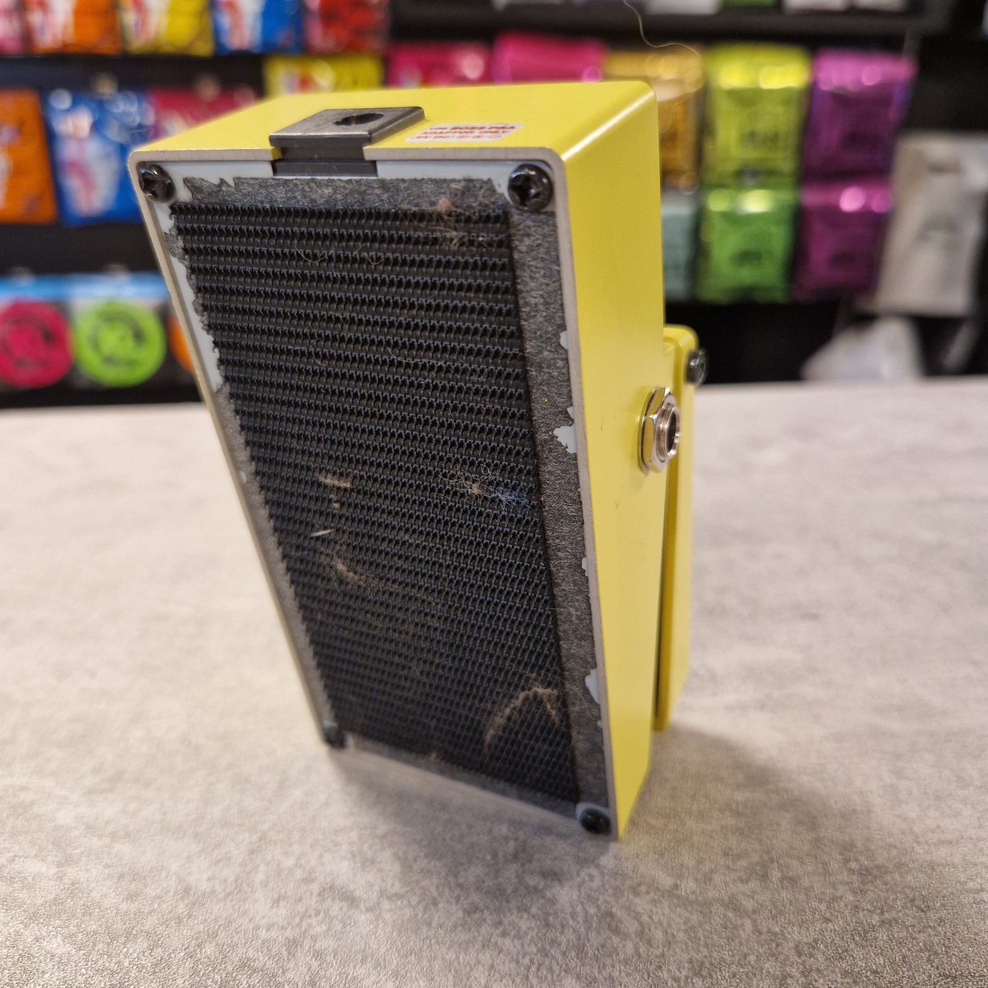 Pre-Owned Boss Yellow electronic device with a black mesh cover on a grey surface, with colourful items in the background.