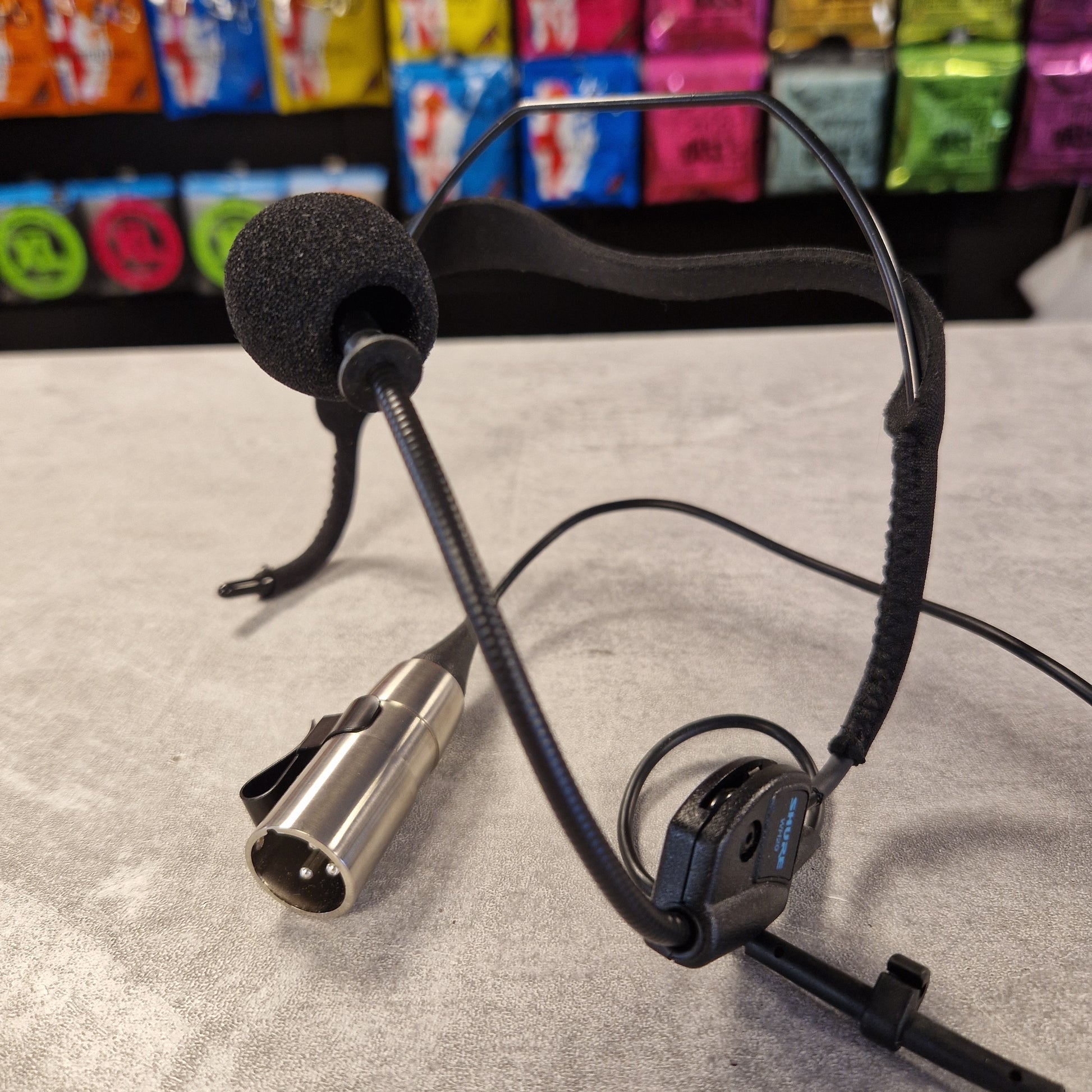 Pre-Owned Shure WH20XLR Headset Microphone - XLR Microphone with cable and metal connector on a textured surface with colourful packaging in the background