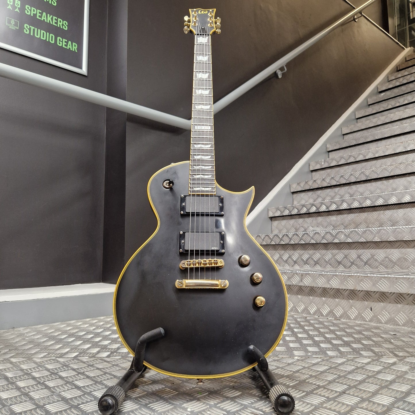 Pre-Owned ESP Black electric guitar with gold hardware on a stand in a room with acoustic panels.