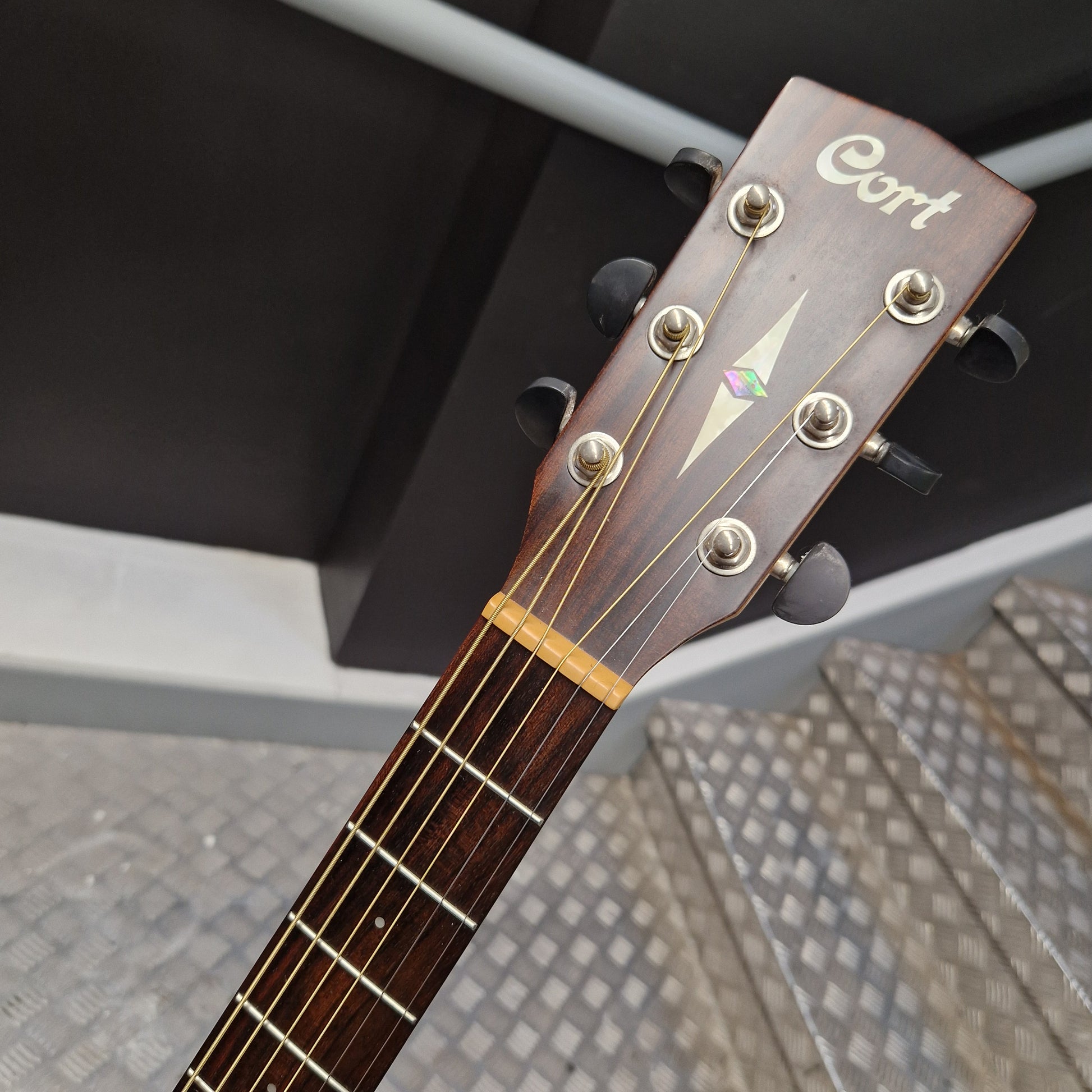 Pre-Owned Cort Close-up of a guitar headstock with 'Cort' branding on a blurred outdoor background.