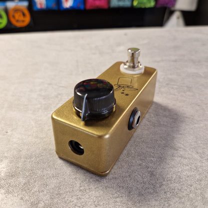Pre-Owned JHS Gold guitar effects pedal on a gray surface with colorful objects in the background