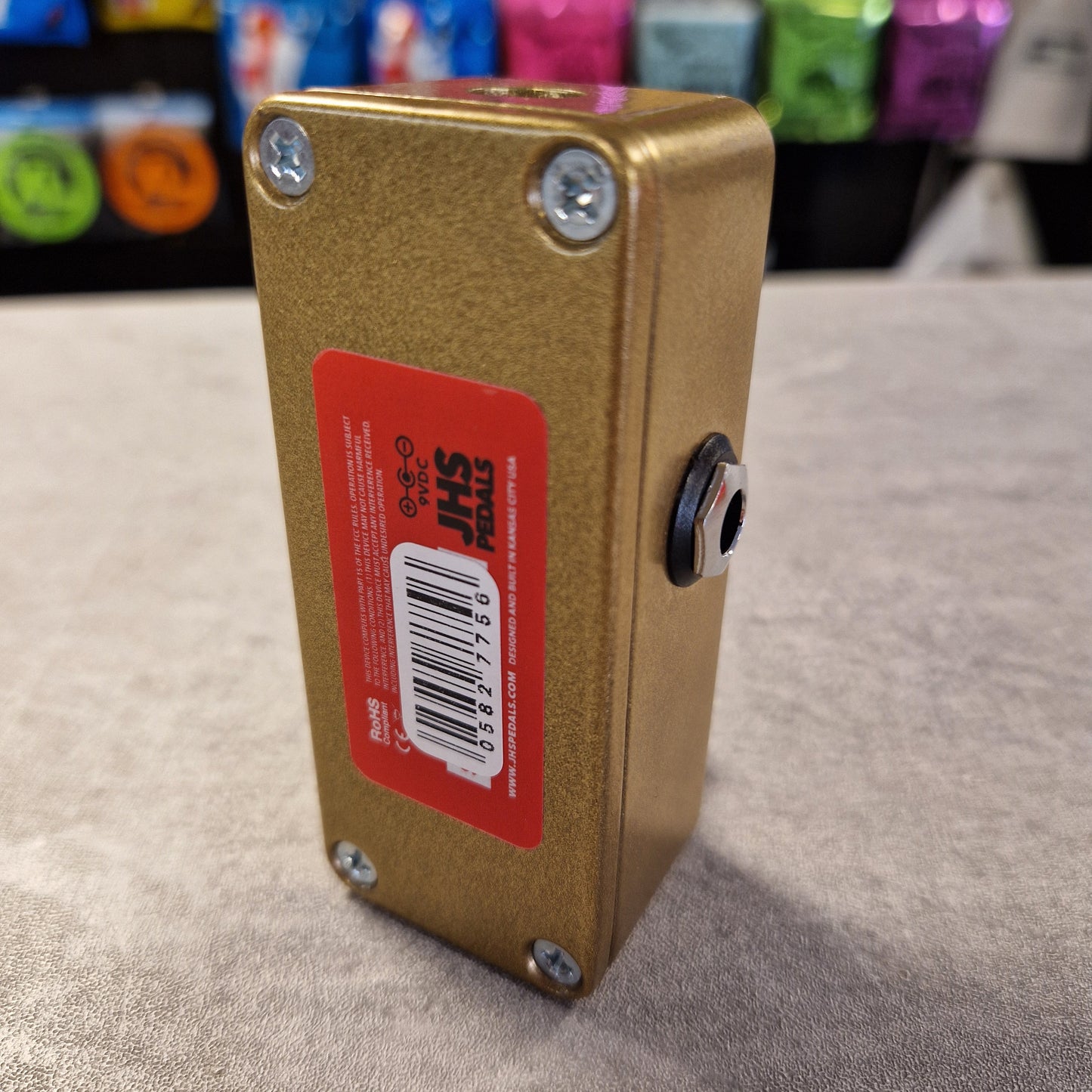 Pre-Owned JHS Gold rectangular box with a red label featuring a barcode on a gray surface.