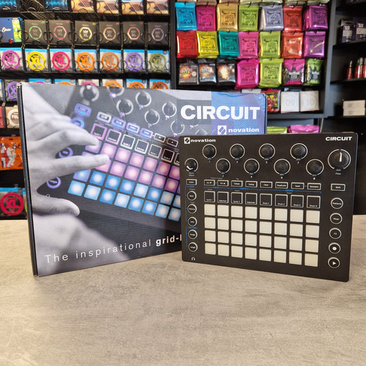 Pre-Owned Novation Music production controller with colorful pads and a book titled 'Circuit' on a wooden surface.