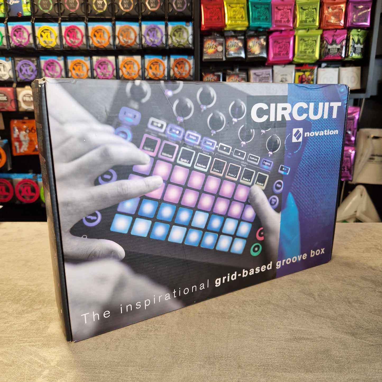 Pre-Owned Novation Novation Circuit music production device packaging on a wooden surface with colorful boxes in the background.