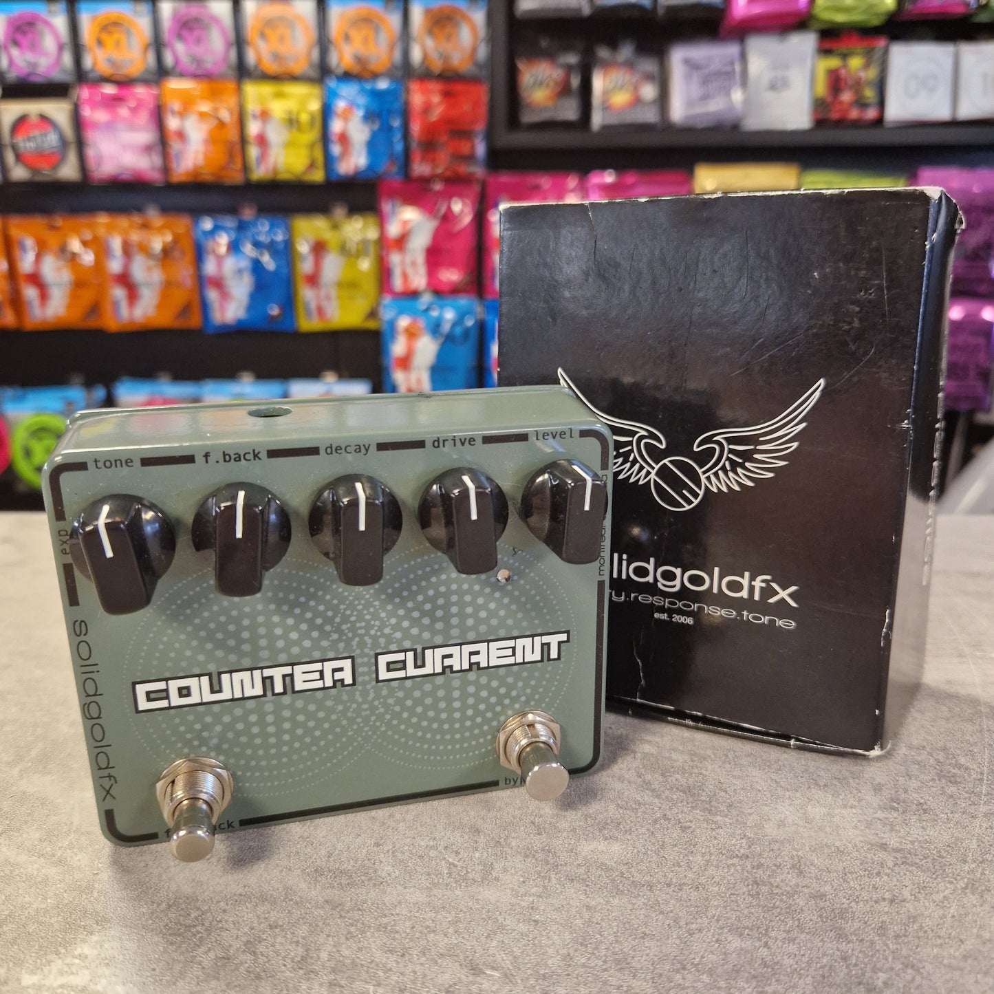 Pre-Owned SolidGoldFx guitar effects pedal on a gray surface with colorful boxes in the background