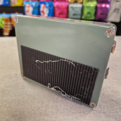 Pre-Owned SolidGoldFx Metal box with a mesh cover on a gray surface, with colorful items in the background.