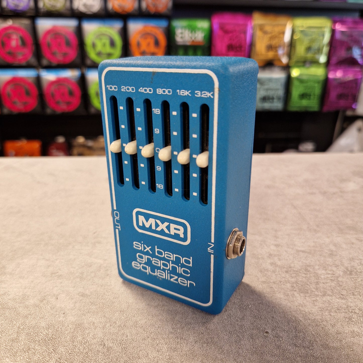 Pre-Owned MXR MX-109 Blue MXR six band graphic equalizer pedal on a gray surface with colorful boxes in the background.