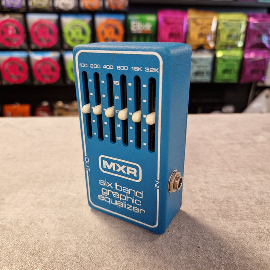 Pre-Owned MXR MX-109 Blue MXR six band graphic equalizer pedal on a gray surface with colorful boxes in the background.