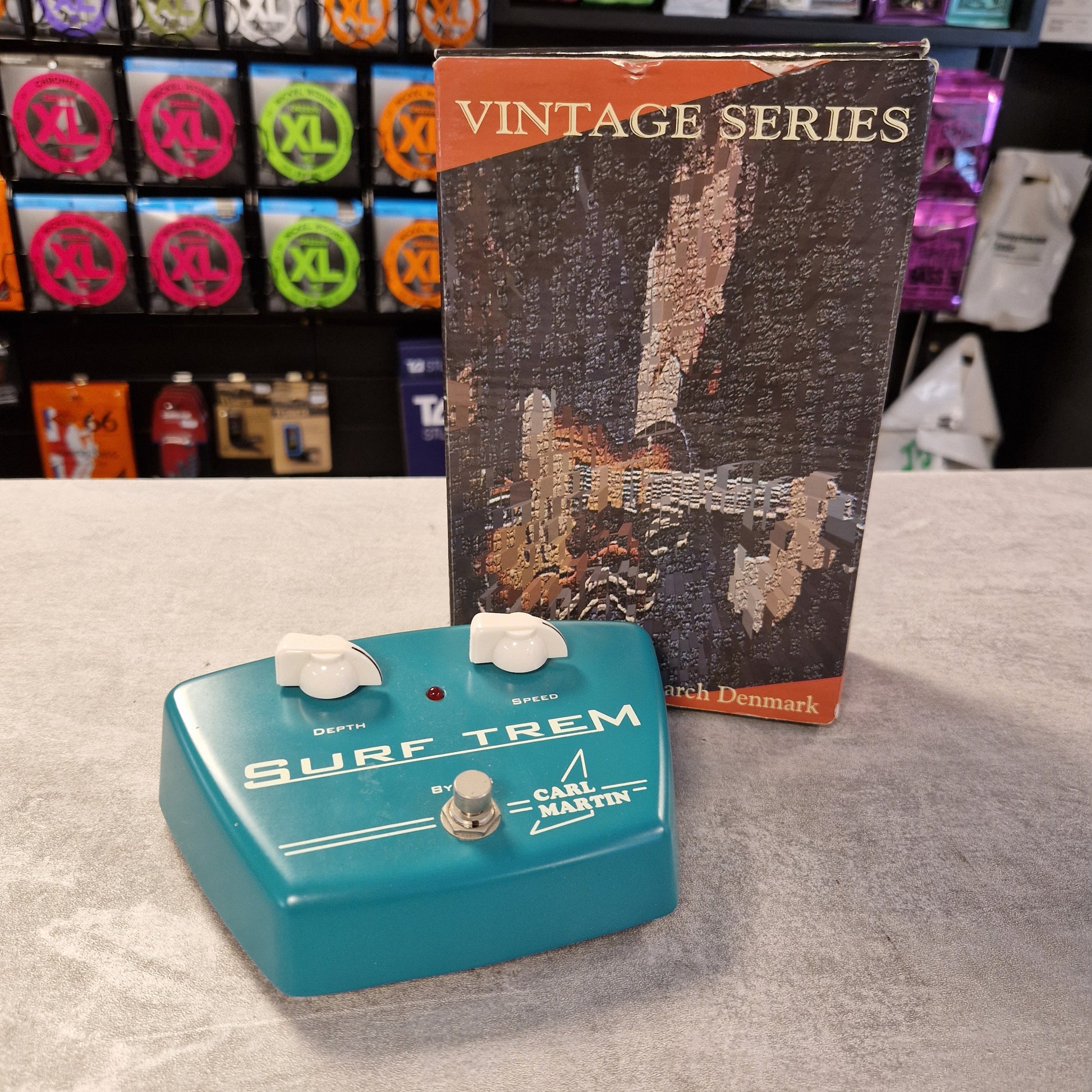 Pre-Owned Carl Martin Surf Trem Blue guitar effects pedal with 'Surf Trem' branding on a gray surface, next to a vintage-style product box.