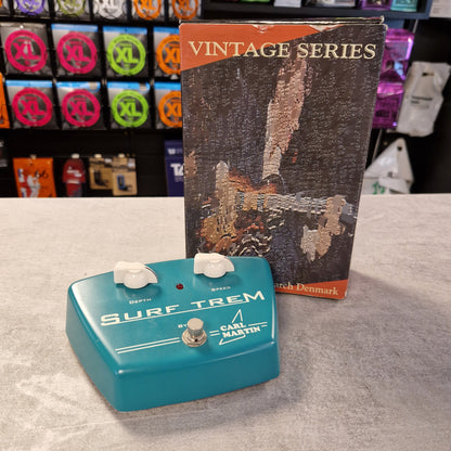 Pre-Owned Carl Martin Surf Trem Blue guitar effects pedal with 'Surf Trem' branding on a gray surface, next to a vintage-style product box.