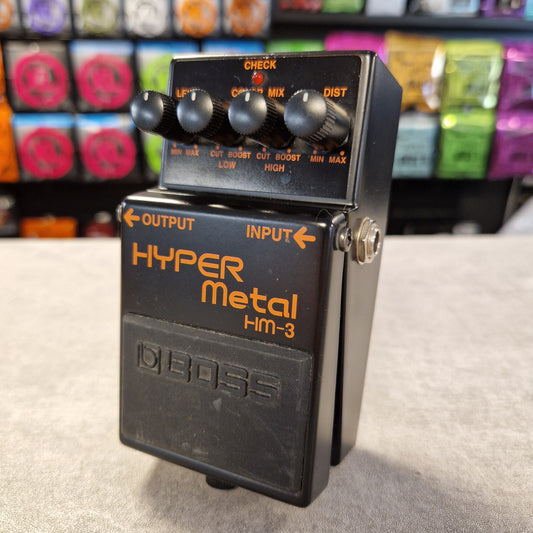 Pre-Owned Boss HM-3 Boss Hyper Metal HM-3 guitar effects pedal on a textured surface with colorful equipment in the background.