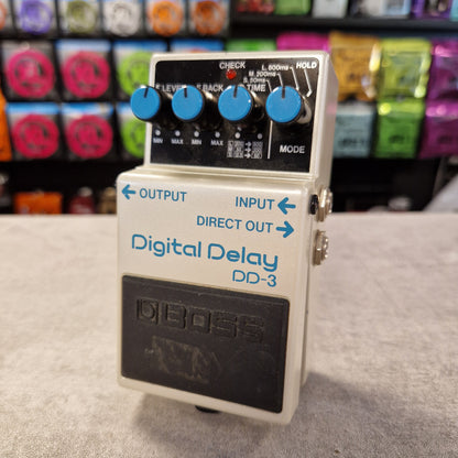 Pre-Owned Boss DD-3 Boss Digital Delay DD-3 guitar effects pedal on a gray surface with colorful equipment in the background.